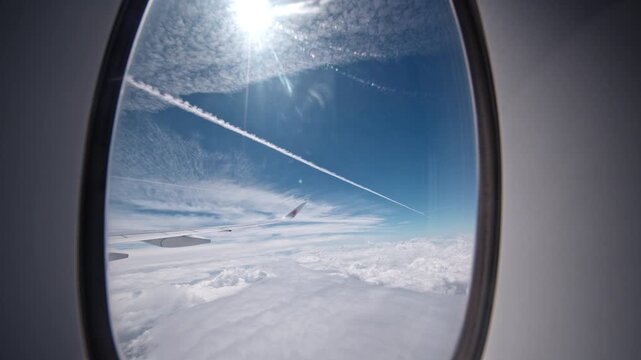 A jet contrail stretches across a clear blue sky, viewed from an airplane window. The sun shines brightly, casting light on the scene. Useful for travel and aviation themes.