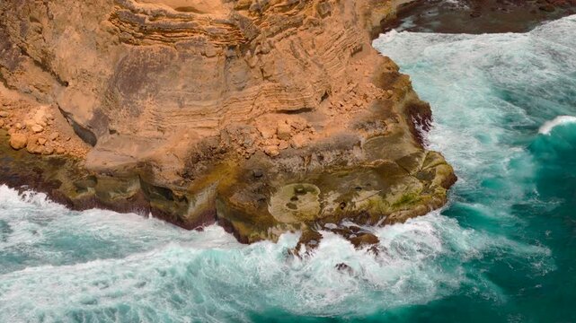 Close ekas drone view shows white ocean waves crashing against a rocky cliff base on wild tropical coast for dramatic nature power geology and shoreline texture themes