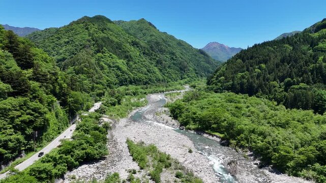 Aerial view of a winding river through green mountains. Clear blue sky above, trees in full leaf. Riverbed shows stones and flowing water. Path runs alongside, barely visible. Scenic view evokes peace