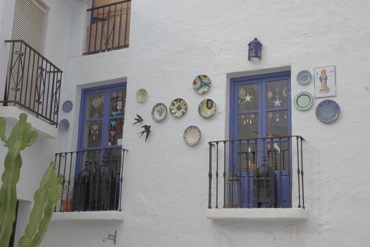 Whitewashed building facade decorated with colorful ceramic plates and metal swallow ornaments &mdash; traditional Andalusian home exterior with purple window frames, iron balconies and cactus, Frigiliana, 
