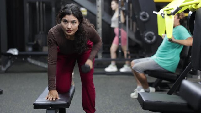 Latina woman standing near a bench in the gym and doing dumbbell rows for back muscles. Motivated woman doing upper body workout for relief arms and back. High quality 4k footage