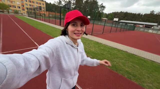 High angle POV selfie of a young active woman in a red cap running on an athletic track, horizontal smartphone video screenshot for fitness blogging and content creation concept
