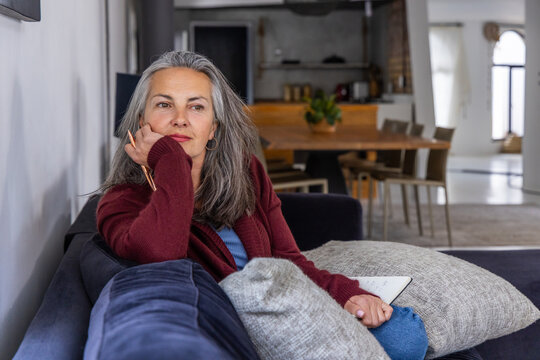 Senior woman sitting on blue sofa at home in maroon cardigan holding pencil, notebook, copy space