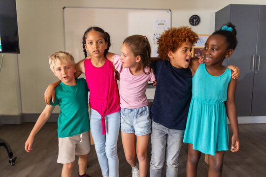 Five children standing with arms around each other in classroom near whiteboard and monitor