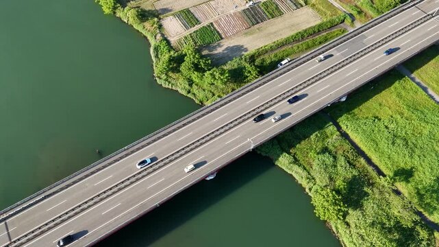 Aerial view of a long bridge crossing a green river. Vehicles move steadily across the asphalt lanes. Side views show lush banks and farmland nearby. No people or animals visible