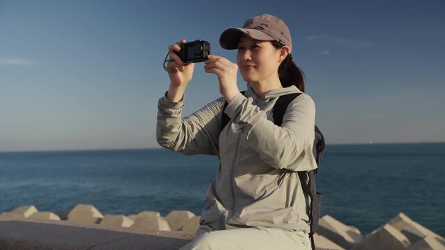 A young female hiker sits on coastal breakwater, checking her compact camera under bright sunny sky by the calm sea.