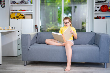 Obraz premium Child relaxing while reading a book at home. Happy kid read book in a home interior. Little reader enjoying a story books at home. Kid reading book and learning in the playroom. Child holding a book.
