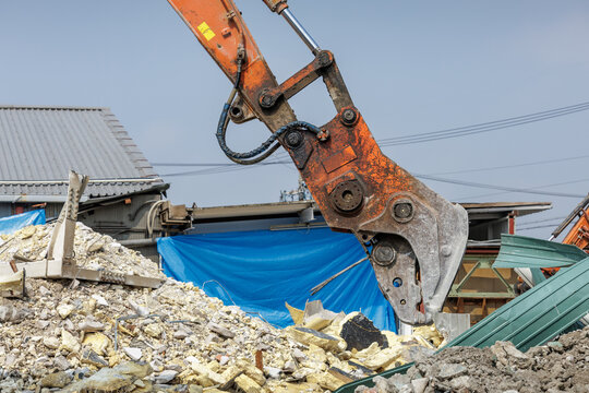 Orange hydraulic excavator crusher arm extends forward over a pile of shattered concrete, metal framing and insulation rubble, readying to further crush debris into manageable pieces.