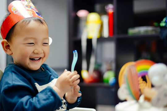 Close-up portrait of a cute 3 year old Chinese boy wearing a birthday hat and smiling happily while eating cake at home, China