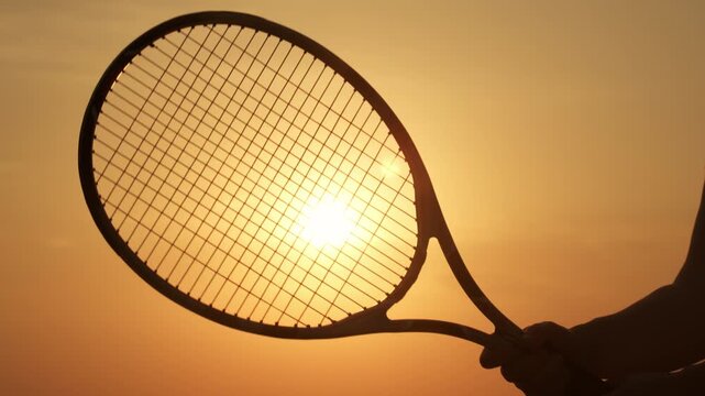 Silhouette of tennis racket in hands of tennis player against background of sun in sunset orange sky close-up