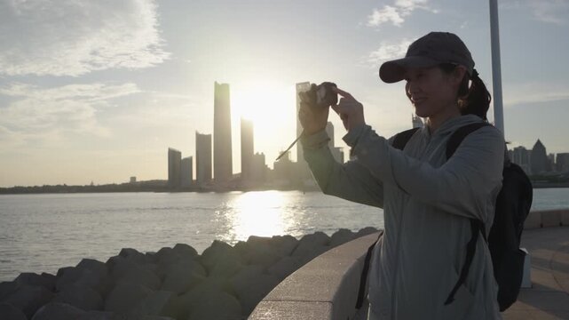 A smiling adult female backpacker walks along a paved urban waterfront promenade at golden hour, with a modern city skyline across the water.