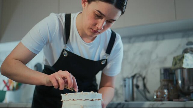 Young woman carefully layers a cake with frosting. She focuses intently, showing patience and care. Kitchen setting suggests home baking or professional dessert preparation