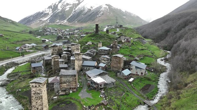Aerial view of the ancient high altitude settlement in Ushguli, showing traditional fortified tower houses, a rushing river fork, and green valley slopes under a dramatic Caucasus mountain ridge.