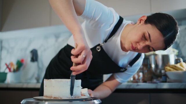 Young female chef carefully cuts a cake. She focuses intently, showing precision and care. The kitchen background shows clean, modern equipment. This footage works well for food, baking