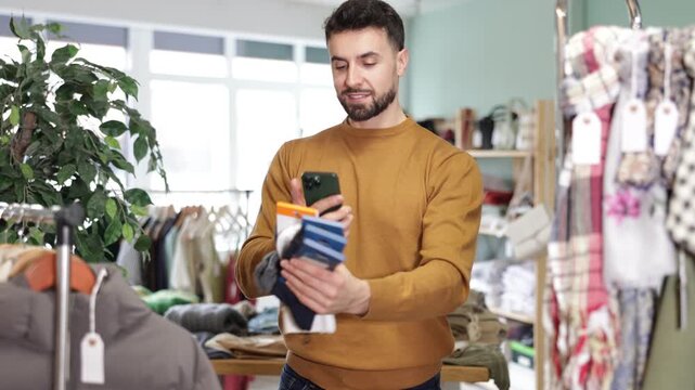 Bearded man stands in a clothing store and scans the barcode of the label on packages of socks. Buyer uses a modern payment method by qr. High quality 4k footage
