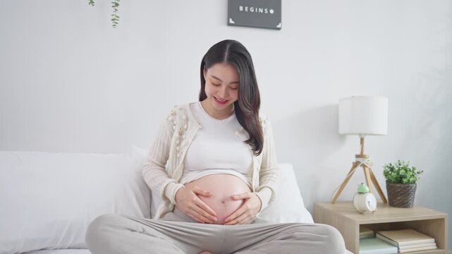 Asian young pregnant woman gently stroking belly while sitting on bed