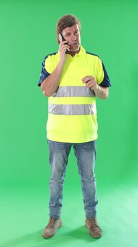 Adult male worker wearing reflective polo shirt stands against green screen, holding smartphone and speaking