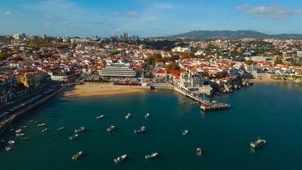 Fototapeta premium Cascais city displaying its vibrant coastal urban landscape with many boats dotting the tranquil water of the bay, a sandy beach, and historical architecture under a bright blue sky