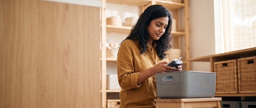 Indian woman organizing home storage using label maker device at home. Smiling South Asian female sorting pantry containers, decluttering closet, lifestyle, productivity, in a modern minimalist room.