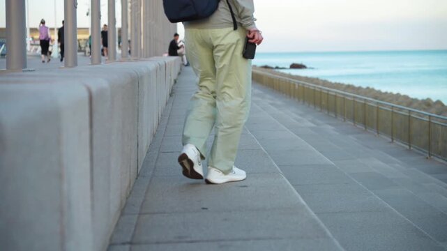 A casual adult traveler with a backpack walks along a paved seaside promenade, with the ocean on one side and architectural columns on the other.