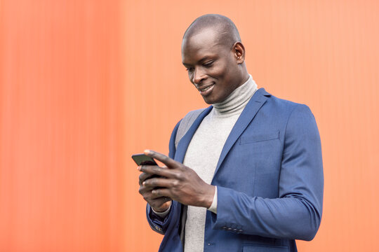 Smiling businessman standing in front of orange wall looking at smartphone