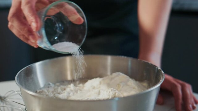 Hand pours water into flour in metal mixing bowl. Close-up shot of baking preparation. Smooth motion, clean kitchen background. Ideal for recipes, cooking tutorials, or food blogs