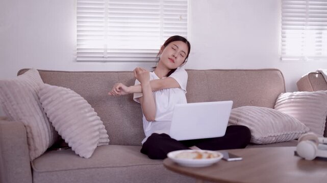 young Asian woman stretching arms relaxing with laptop computer on sofa at home after remote work morning routine lifestyle wellness break comfortable living