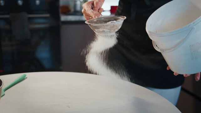 Person sifting flour into bowl, kitchen background. White bucket held in hand, flour dust flies. Action focused, hands steady, motion clear. Use for baking, cooking, food prep, or culinary projects