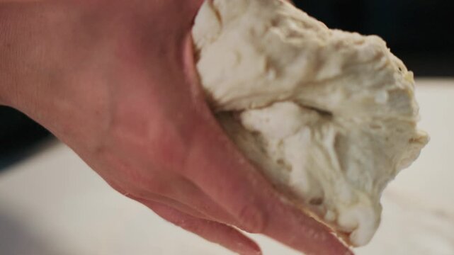 Close-up of hands kneading white dough on a clean surface. Fingers press into sticky, elastic dough, showing texture and effort. Scene suggests home cooking, artisan bread, or culinary tutorial