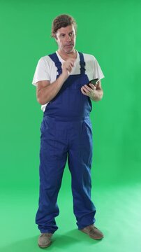 Middle-aged construction man stands in overalls and holds a smartphone in his hands against the background of chromakey. Handyman in overalls responds to customer messages on green background. High