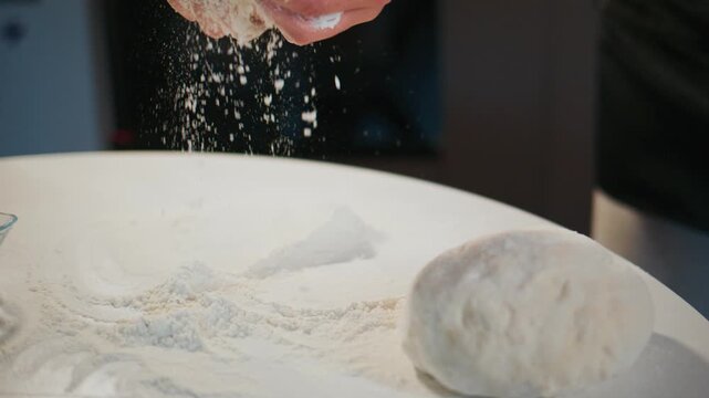 Hands knead white dough on floured table. Flour dusts air as hands work. Scene shows baking preparation. Ideal for recipes, food blogs, or cooking tutorials. Focus on tactile, hands-on baking process