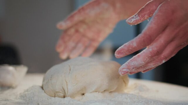 Close-up of hands gently placing dough on floured surface. Hands lightly dusted with flour, showing careful touch. Dough appears soft and pliable, resting on white powder. Ideal for cooking tutorials