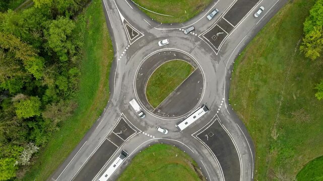 Aerial drone slow zoom-in on busy roundabout with multiple trucks and trailers navigating intersection alongside cars. Heavy freight traffic at logistical junction with green surroundings from above.