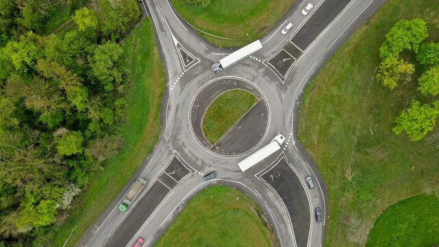 Static aerial drone view of roundabout with vehicle density and minor congestion as trucks and buses navigate intersection. Traffic flow and queuing behavior visible from overhead with green