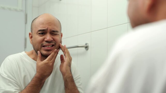 Concerned middle-aged man touching cheek and suffering toothache while looking in mirror at home. Dental pain, oral health problem, healthy aging, men's wellness and self-care concept.

