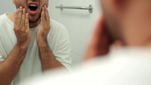 Concerned middle-aged man touching cheek and suffering toothache while looking in mirror at home. Dental pain, oral health problem, healthy aging, men's wellness and self-care concept.
