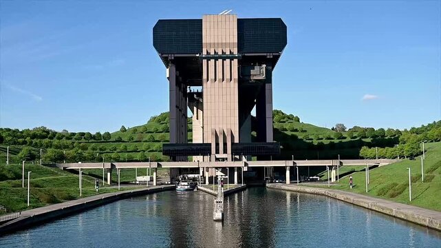 Static wide view of cargo barge carefully entering canal boat lift lock chamber with concrete towers on both sides and green hillside behind. Vessel navigating into caisson with calm water and summer