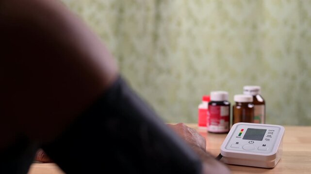Male adult patient checks blood pressure level utilizing small digital monitor device placed atop brown wooden table surface, near multiple plastic medicine bottles inside soft home indoor setting.