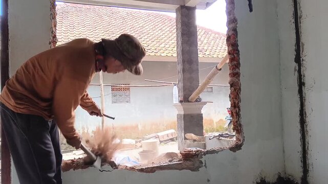 Asian construction worker breaking down a brick wall using manual tools. Hardworking laborer performing demolition tasks for residential building project.