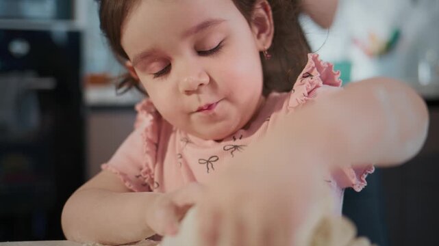Young girl kneads dough with focused attention. She wears pink ruffled top, eyes intent on task. Hands move gently, shaping soft white mixture. Warm kitchen light highlights her concentration
