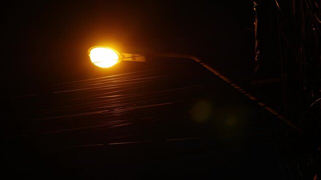 Dramatic low angle night view of tangled electric power cable wires on utility pole with glowing orange street lamp against dark blue sky