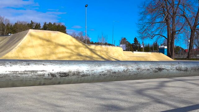 Empty concrete skatepark with various ramps and a metal grind rail in the foreground on a bright sunny day. The clear blue sky and tree shadows create a serene, yet dynamic, urban sports scene