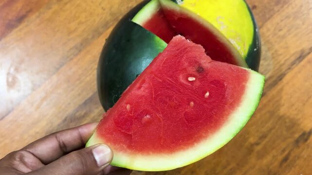 fresh red watermelon slice held by hand with ripe watermelon on wooden table background