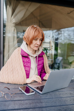 Focused red haired woman typing on laptop near glass cabin wall