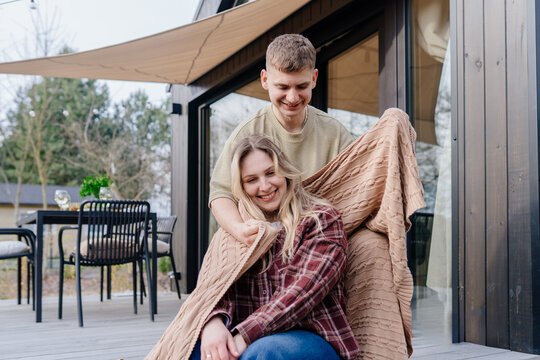 Smiling young white couple sharing blanket outside wooden cabin