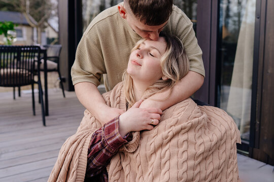 Young white man kissing girlfriend on cabin terrace under blanket