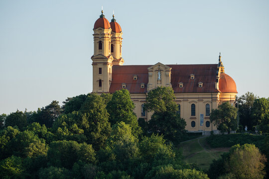 Historic baroque pilgrimage church (Wallfahrtskirche Sch&ouml;nenberg) with twin towers and a red-tiled roof, surrounded by lush green trees at golden hour. Architectural landmark in Bavaria, Germany.