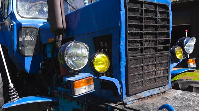 Close up view of an old blue farm tractor's front end, showing its rustic grille, round headlights, and exposed engine components, representing vintage agricultural machinery and rural life