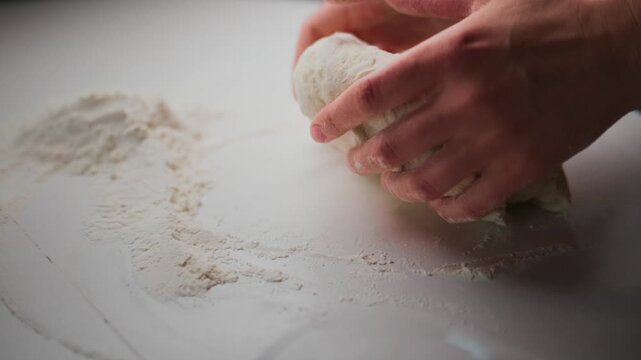 Hands knead white dough on floured surface. Close-up shot shows flour dusting work area. Person focuses intently on shaping dough. Ideal for cooking, baking, or recipe tutorials