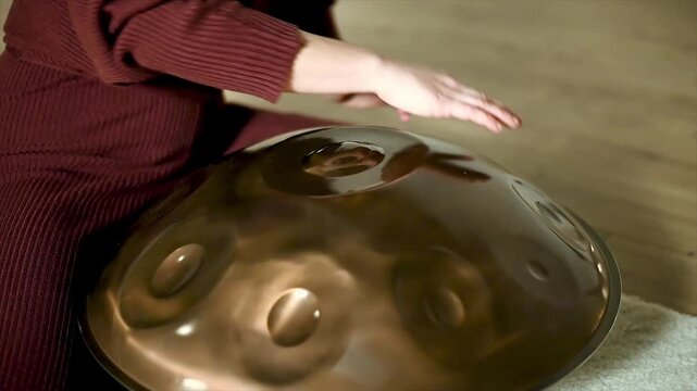 Static close-up of handpan tone field dimples with hands gently striking steel surface during meditation sound healing session. Warm bronze reflections and resonant vibration on handcrafted
